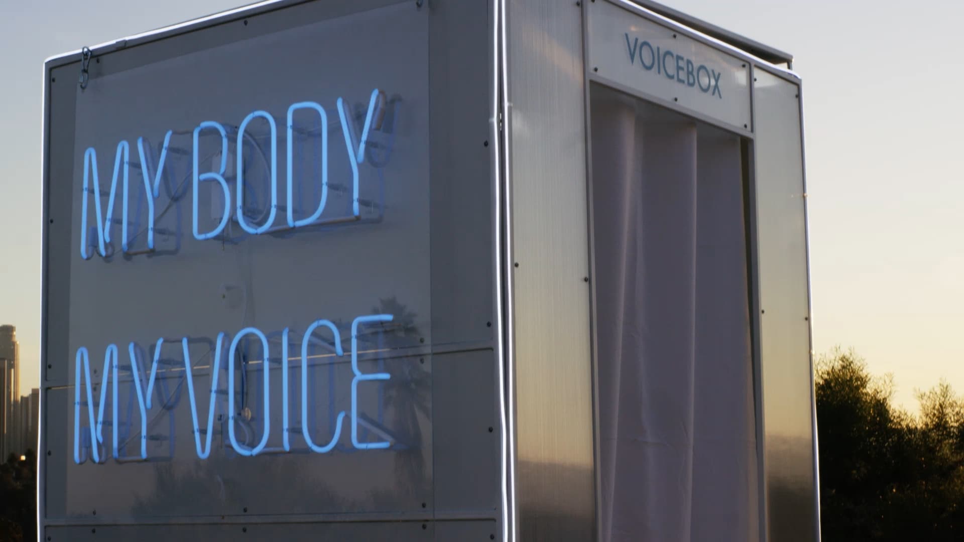 Image of the VoiceBox, in the park, with a glowing neon sign which states, my body my voice.